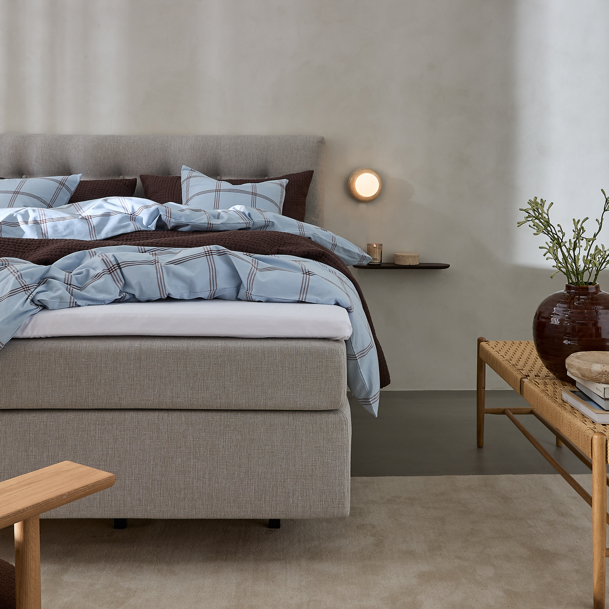 Redt seng i lysegråt med blåternet sengetøj og brun quilt, væglampe og lille hylde med lys, bænk med vase og grønne grene i et roligt soveværelse.