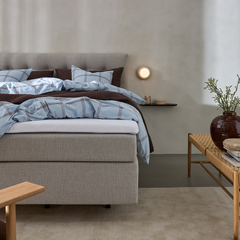 Redt seng i lysegråt med blåternet sengetøj og brun quilt, væglampe og lille hylde med lys, bænk med vase og grønne grene i et roligt soveværelse.