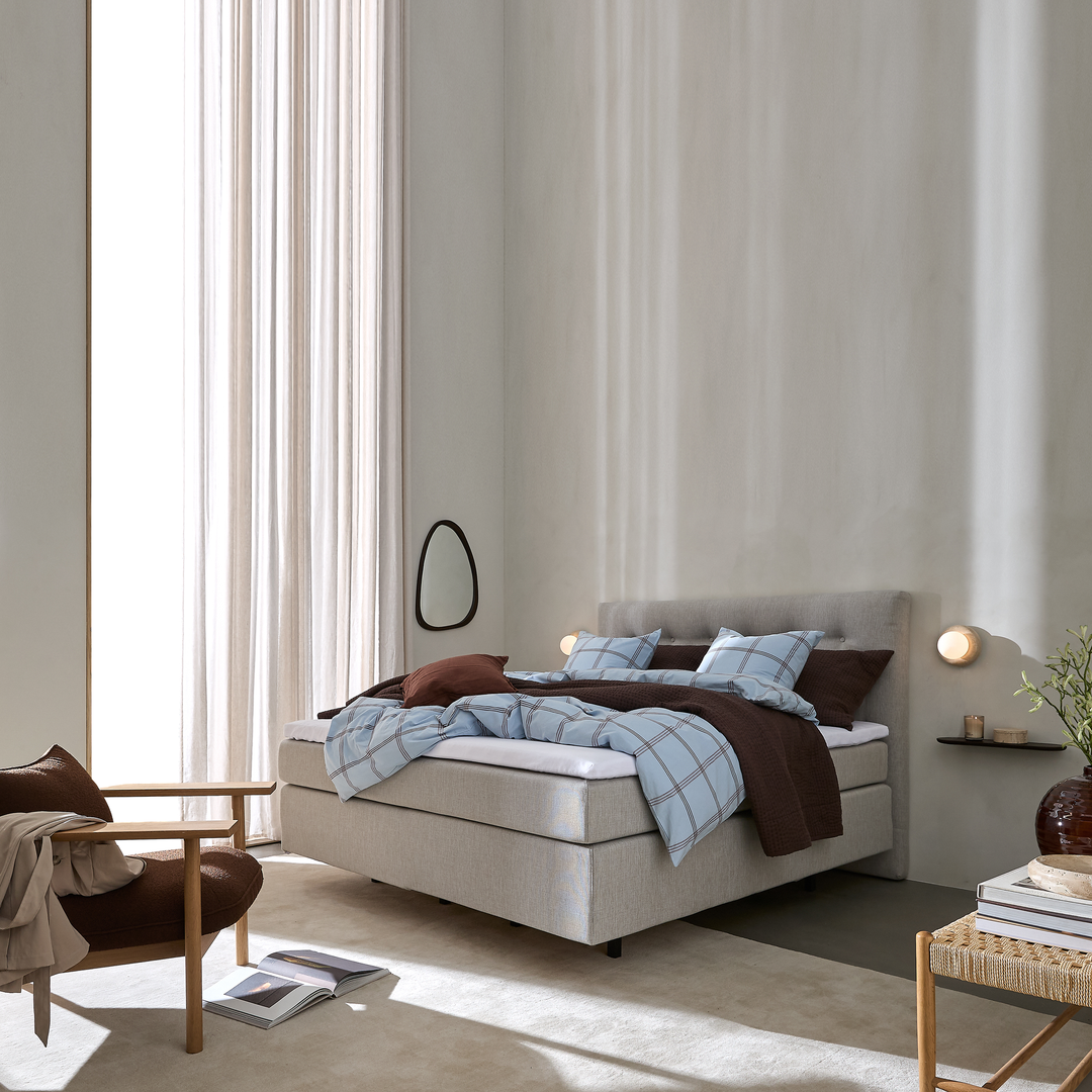 Lyst, minimalistisk soveværelse med beige polstret seng, blårutinet sengetøj og brun plaid; væglamper, spejl og hylde med lys, lænestol og lette gardiner i dagslys.
