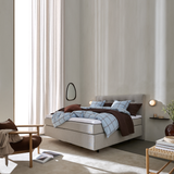 Lyst, minimalistisk soveværelse med beige polstret seng, blårutinet sengetøj og brun plaid; væglamper, spejl og hylde med lys, lænestol og lette gardiner i dagslys.