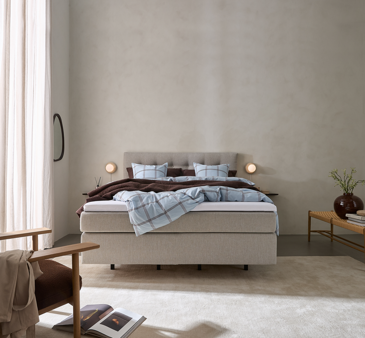 Lyst og roligt soveværelse med beige kontinentalseng, blåternet sengetøj, brun plaid, to små væglamper og bænk med vase og grønne grene.