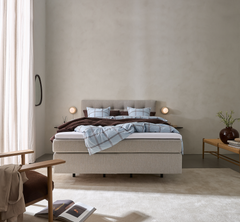 Lyst og roligt soveværelse med beige kontinentalseng, blåternet sengetøj, brun plaid, to små væglamper og bænk med vase og grønne grene.