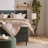 Hyggeligt soveværelse med grå polstret seng, beige sengetøj og plaid, sengebord med lampe og bøger, hylde med dekor og hængeplante samt grøn puf.