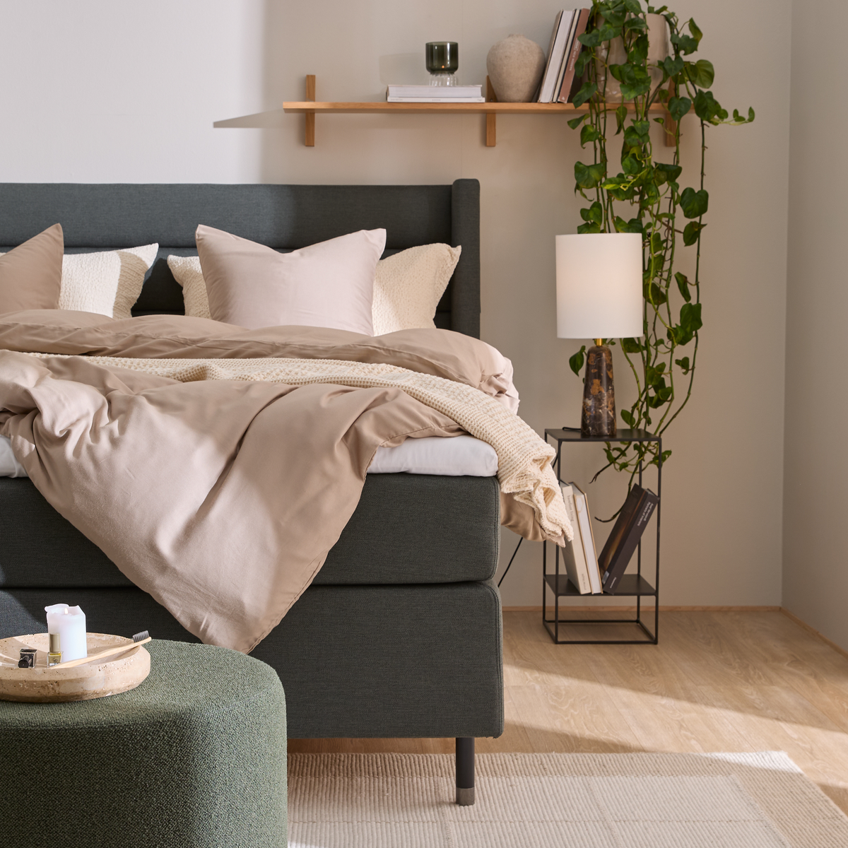 Hyggeligt soveværelse med grå polstret seng, beige sengetøj og plaid, sengebord med lampe og bøger, hylde med dekor og hængeplante samt grøn puf.