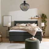 Soveværelse med mørkegrøn seng med beige sengetøj, to bordlamper, sort trådpendel over sengen, væghylde med bøger og hængende plante, grøn puff og lyst tæppe.