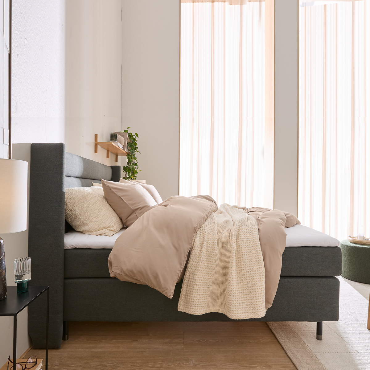 Lyst soveværelse med grå polstret seng, beige dynebetræk, strikket plaid og lyse gardiner i dagslys.