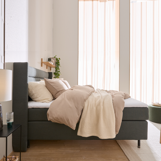 Lyst soveværelse med grå polstret seng, beige dynebetræk, strikket plaid og lyse gardiner i dagslys.