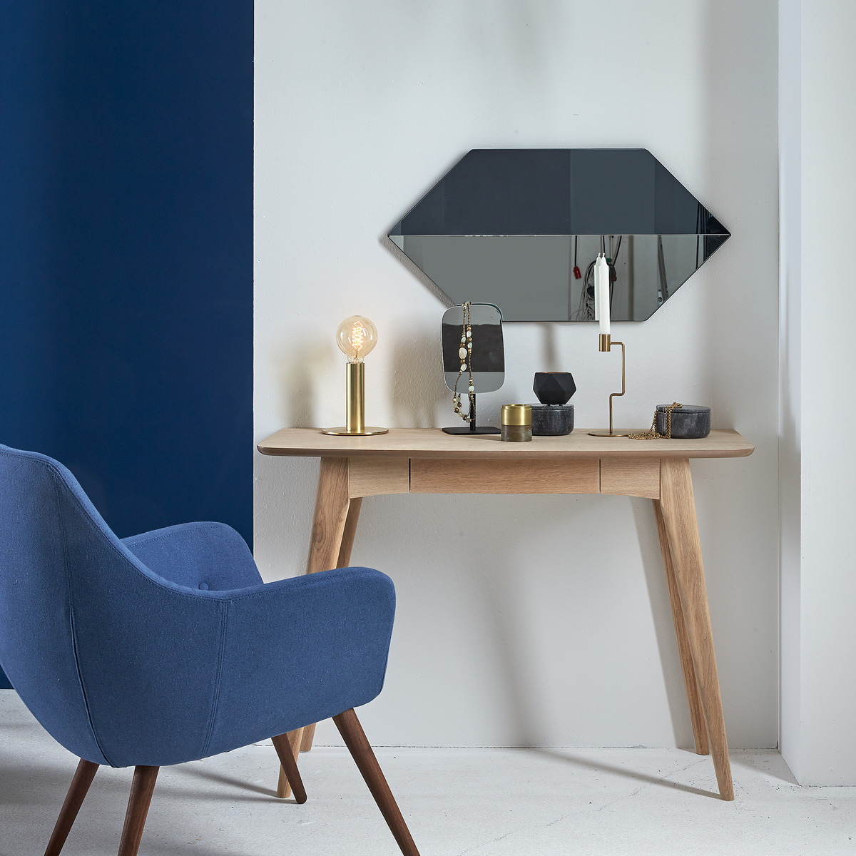 Lysegrå væg med blå accent, en træ-konsol med geometrisk vægspejl over, messingbordlampe med pære, lysestage og små dekorationer; blå lænestol i forgrunden.