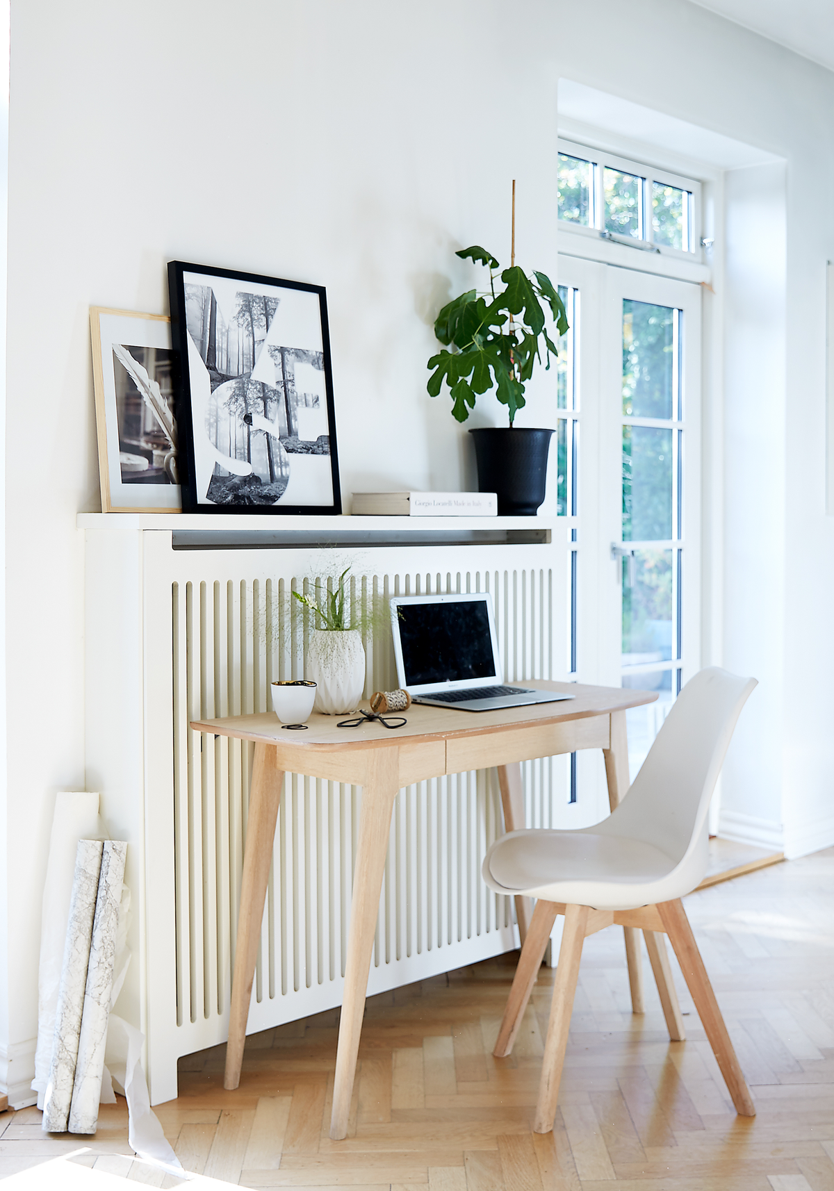 Lyst skandinavisk arbejdsområde med lyst træbord, hvid stol, laptop, grøn potteplante og YES‑kunst foran vinduer i dagslys.