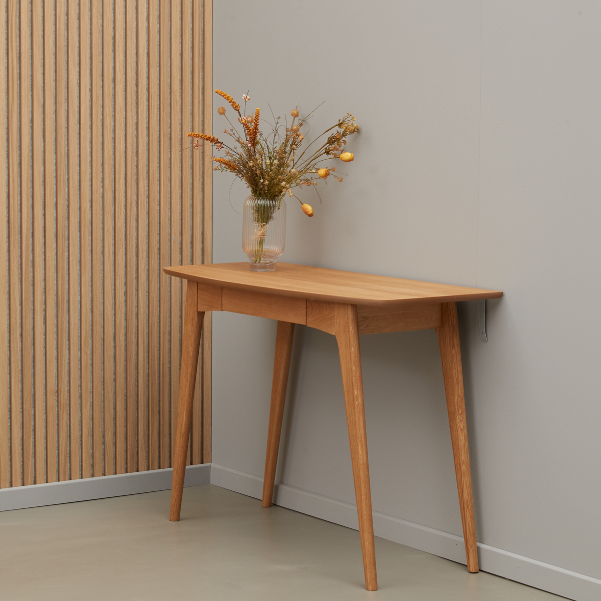 Konsolbord i lys eg med slanke ben og en glasvase med tørrede blomster, mod grå væg og rillet træpanel.