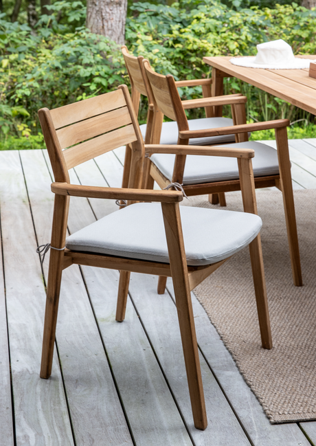 Havestole i teak med lysegrå hynder på en træterrasse ved siden af et matchende bord, grønne planter i baggrunden.