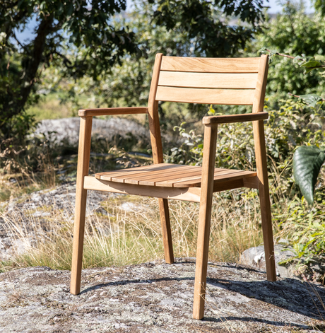 Træstol med armlæn i teak placeret udendørs på en klippe i et solrigt, grønt landskab.