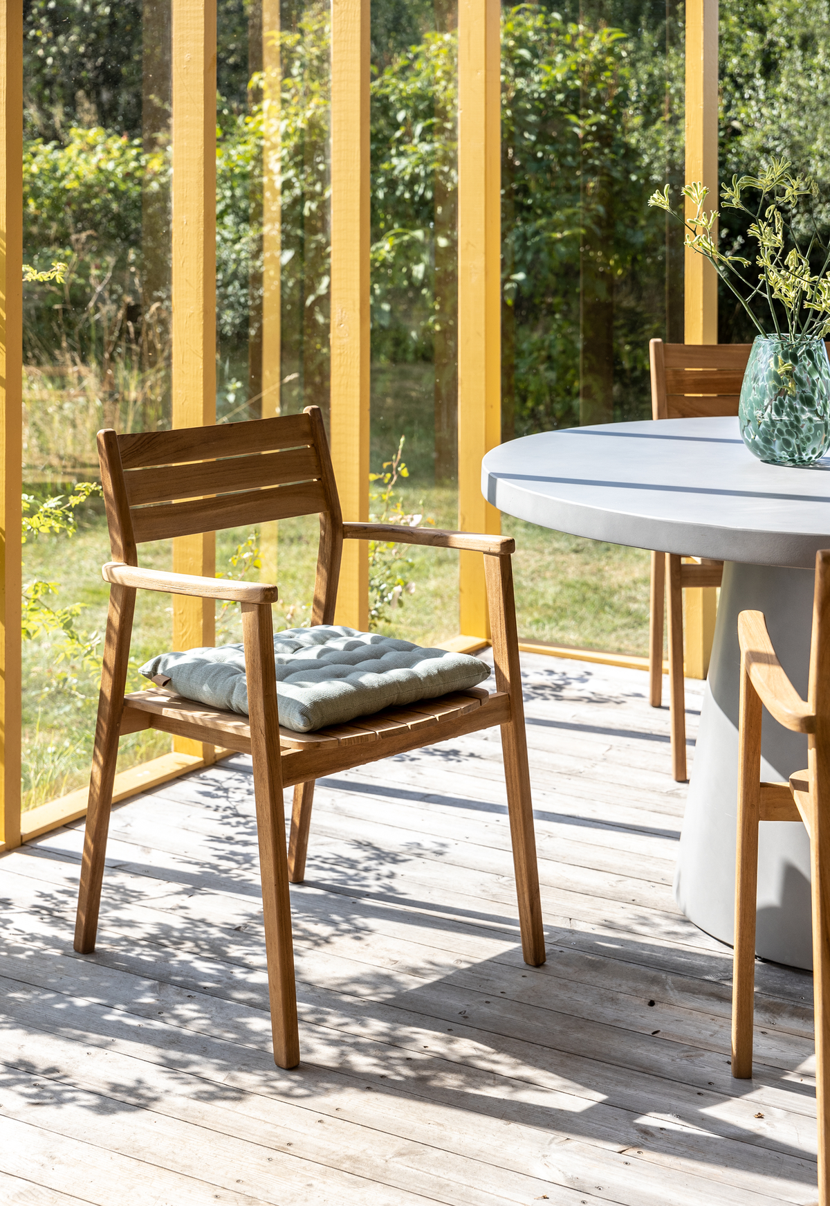 Træstol med armlæn og lysgrå sædepude ved rundt betonbord på solrig terrasse med gule glasrammer og vase med grønne grene.