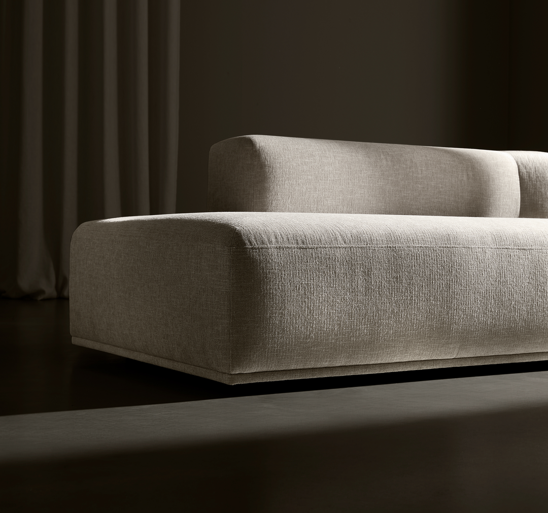 Beigefarvet, lav moderne modulsofa i tekstureret stof mod mørk baggrund med draperet gardin, sideview og dramatisk belysning.