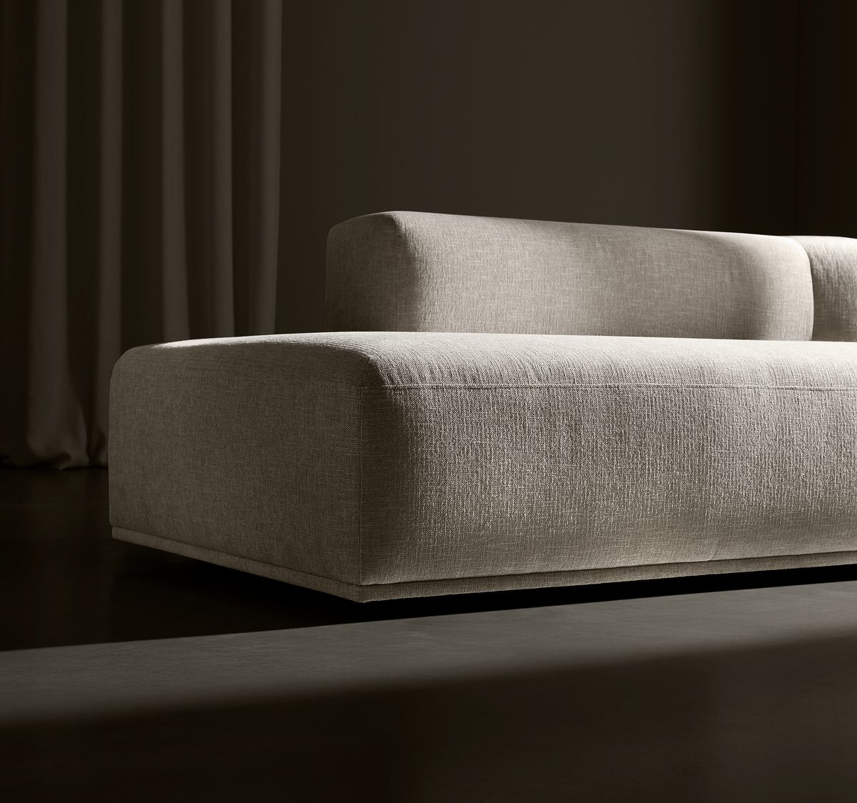 Beigefarvet, lav moderne modulsofa i tekstureret stof mod mørk baggrund med draperet gardin, sideview og dramatisk belysning.