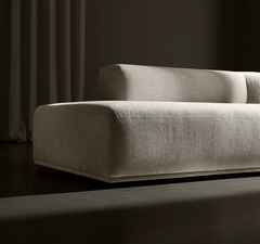 Beigefarvet, lav moderne modulsofa i tekstureret stof mod mørk baggrund med draperet gardin, sideview og dramatisk belysning.