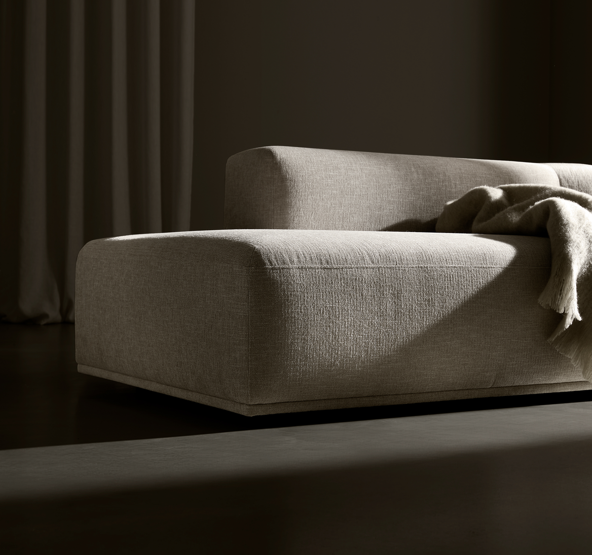 Lav, moderne sofa i beige stof med afrundede kanter og et blødt plaid over armlænet, oplyst af dramatisk sidelys.