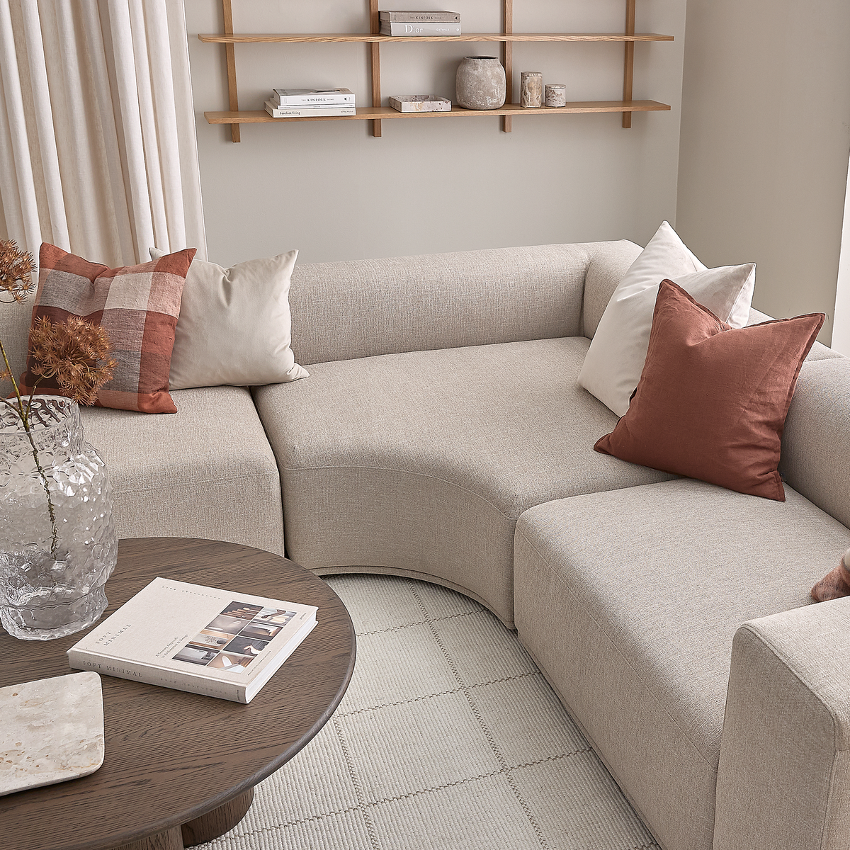 Beige modulsofa med buet hjørne og rust- samt hvide puder, rundt sofabord i mørkt træ med bog og glasvase, åbne hylder med keramik og bøger, lyse gardiner og tæppe i en skandinavisk stue.
