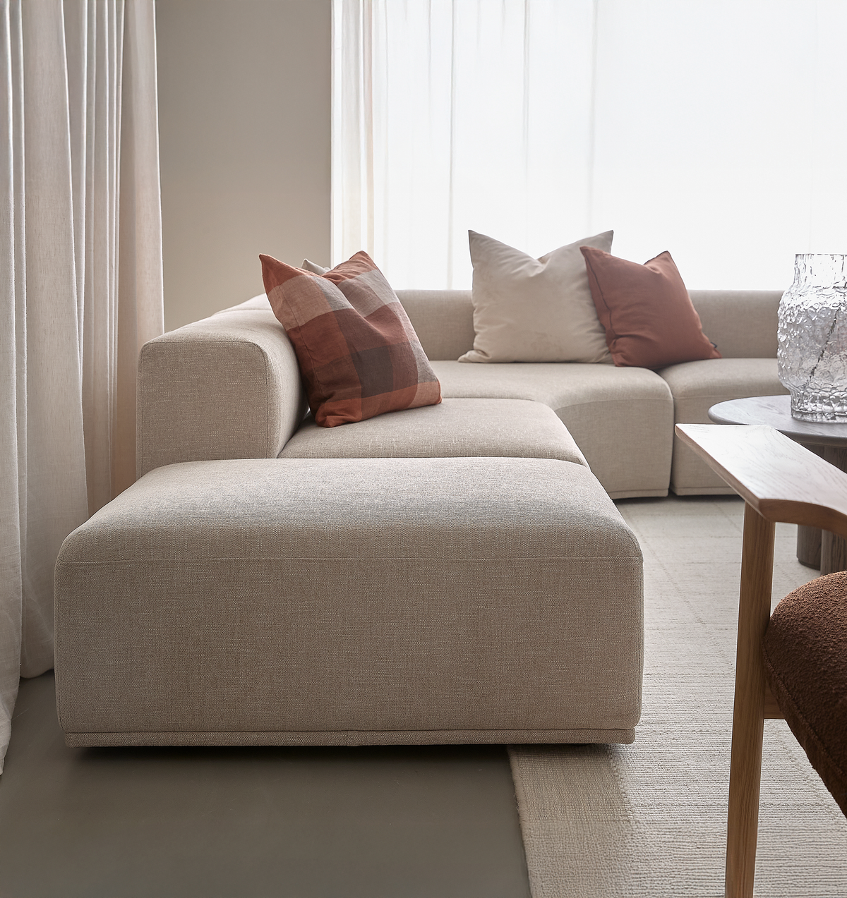 Beige modulsofa i stue med puder i terrakotta og creme, lyse gardiner og træbord i blødt dagslys.