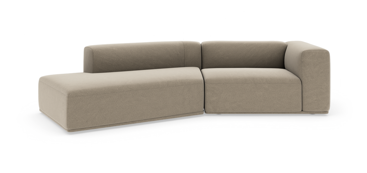 Beige modulsofa i L‑form med venstre chaiselong, lav ryg og brede bløde armlæn, frilagt mod hvid baggrund.