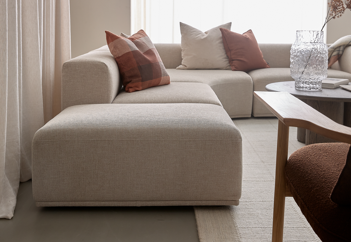 Beige modulsofa med rustfarvede og lyse puder i en lys stue, rundt sofabord med struktureret glasvase og bøger, gardiner og neutralt tæppe.