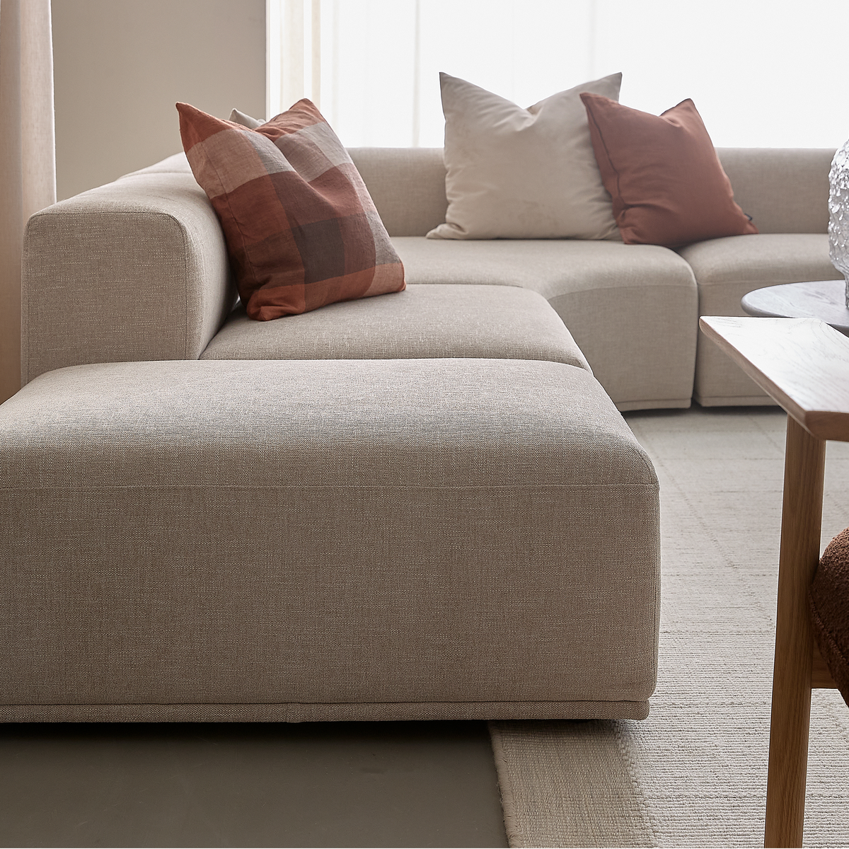 Beige modulsofa med rust- og cremefarvede puder ved siden af et lille træbord i en lys stue.