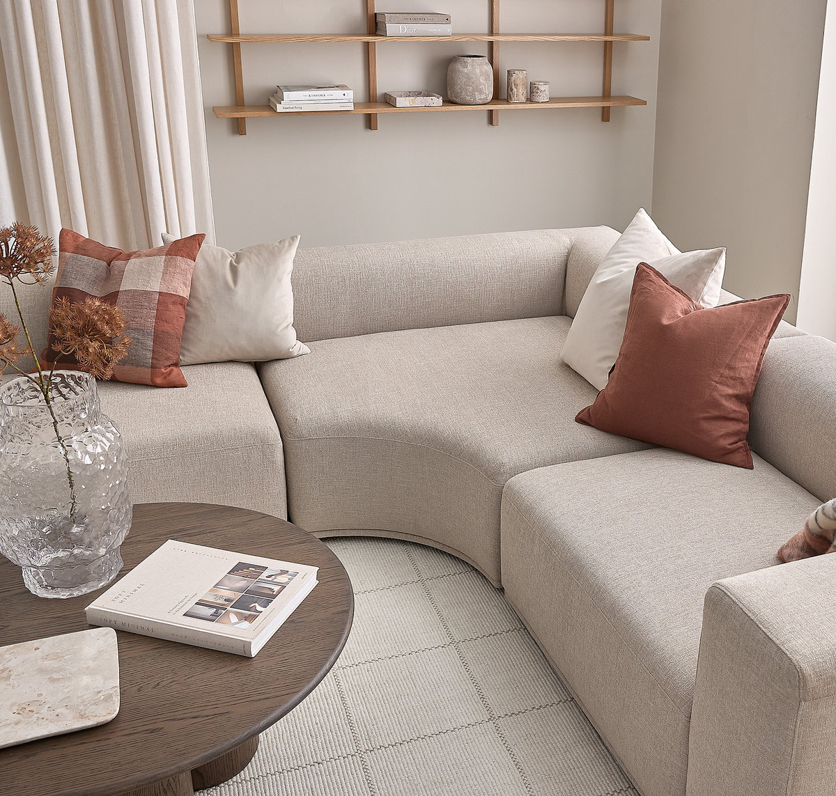 Lys beige modulsofa med bløde kurver og puder i rust og hvid ved siden af et rundt træbord med bogen ”Soft Minimal”, en lys stenplade og en klar struktureret vase med tørrede grene; i baggrunden lyse gardiner og enkle hylder med bøger og keramik.