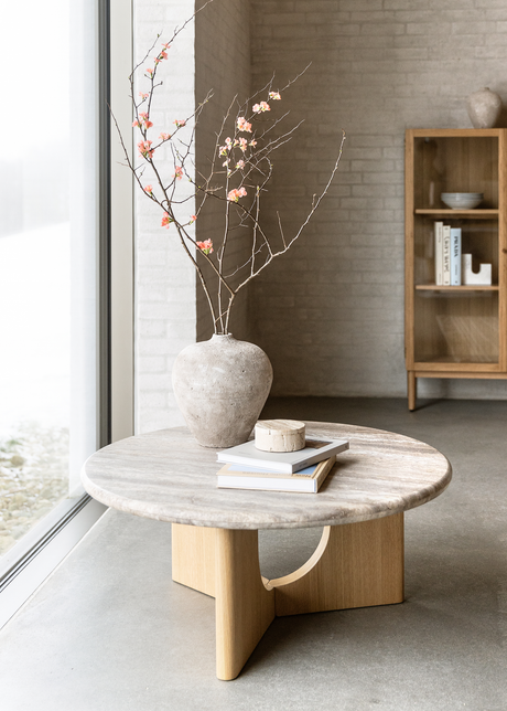 Rundt sofabord i lys sten og træ med en vase med blomstrende grene og stablede bøger ved et stort vindue i en lys stue.
