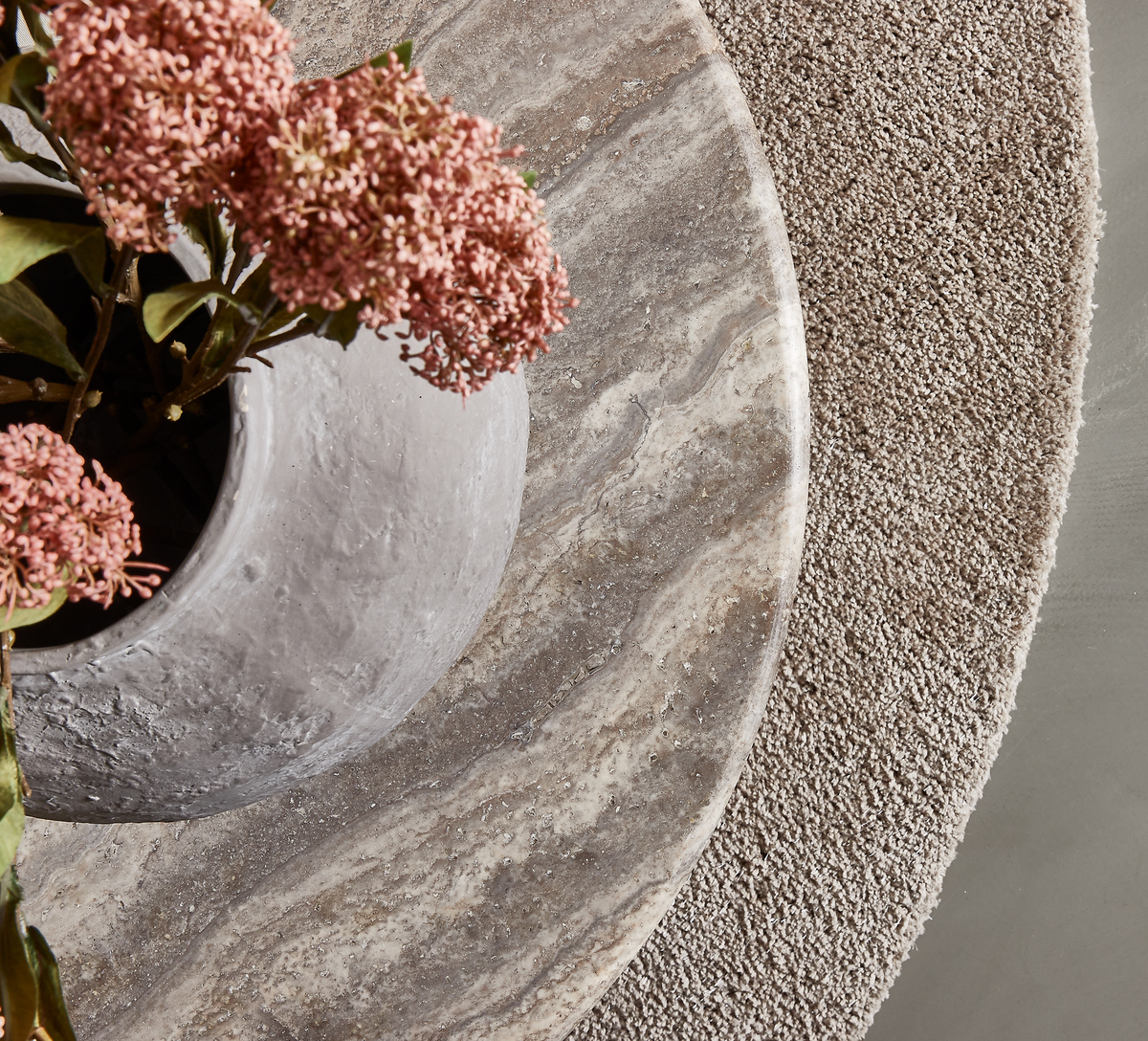 Nærbillede af et rundt stenbord med grå vase og lyserøde blomster på et beige, rundt tekstureret tæppe.