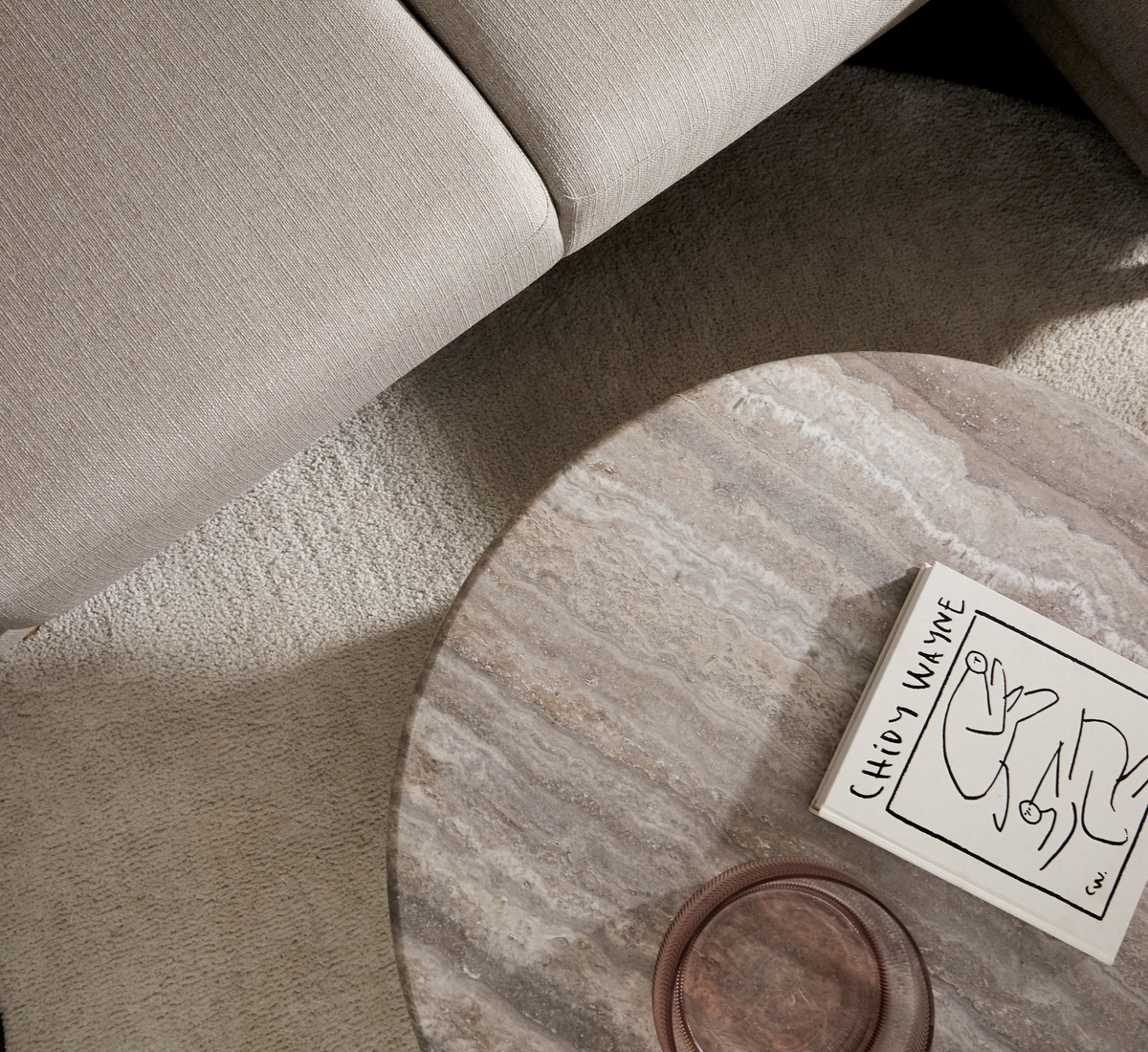 Rundt sofabord i sten med glasfad og bog 'CHIDY WAYNE' på beige tæppe ved siden af lys sofa, set oppefra.