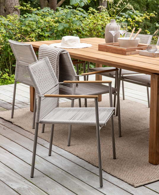 Udendørs spiseområde på en terrasse: træbord med grå rebstole, beige tæppe og en bakke med vase, glas og skål; en hvid hat ligger på bordet omgivet af grønne planter.